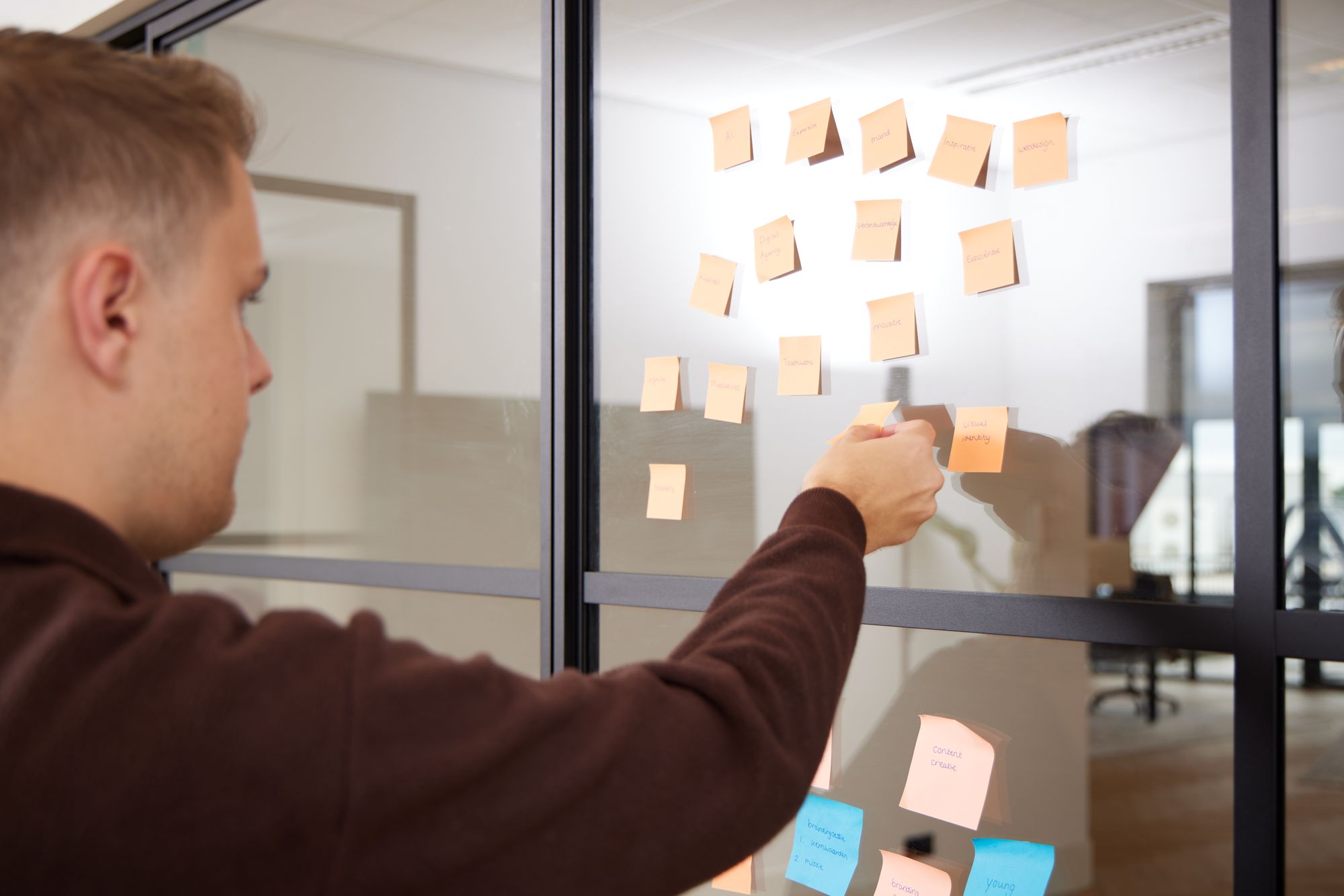 Persoon met donkere trui plakt oranje en blauwe post-its op glazen wand in modern kantoor