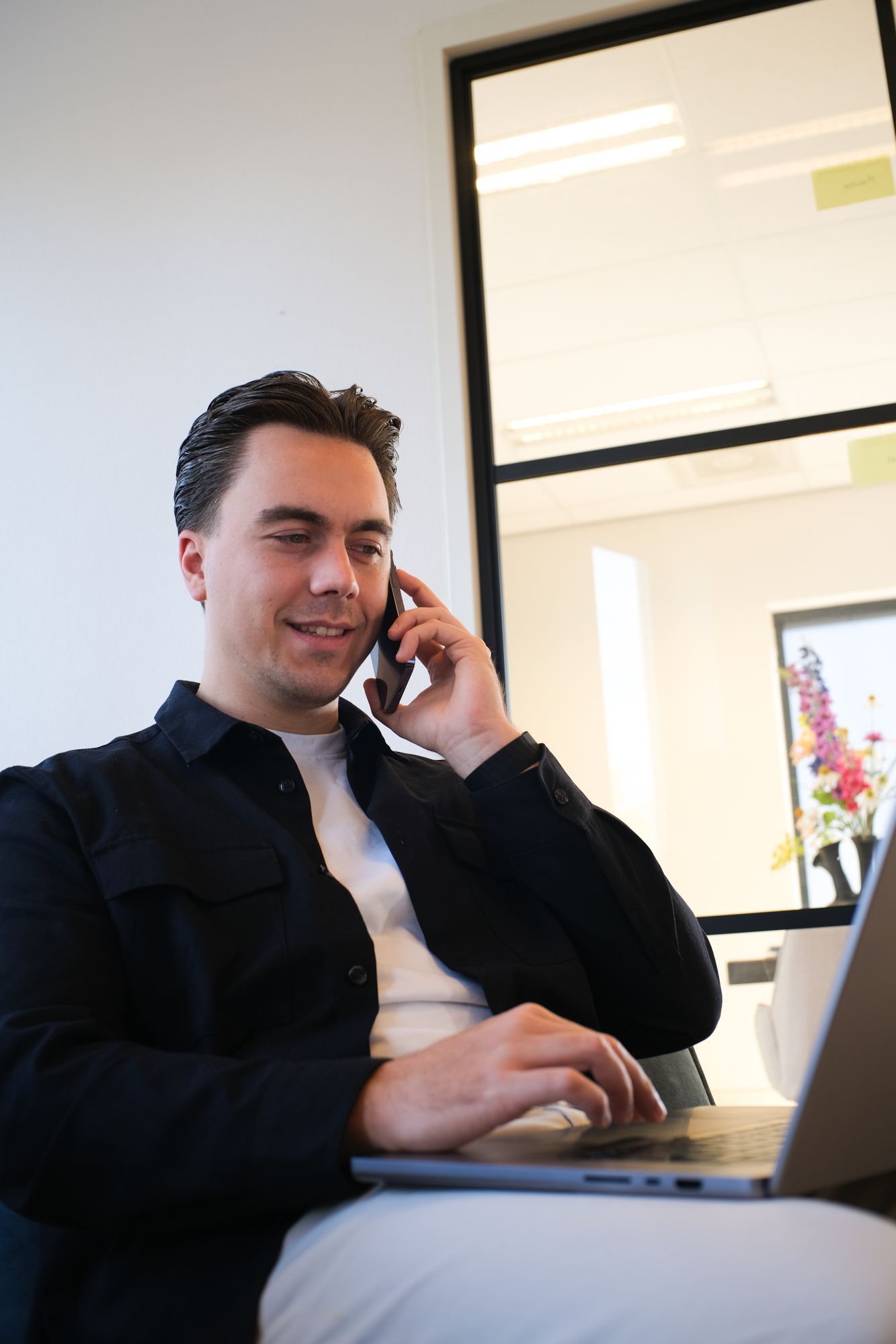 Jonge man zit aan bureau en belt met rechterhand, typt op laptop met linkerhand