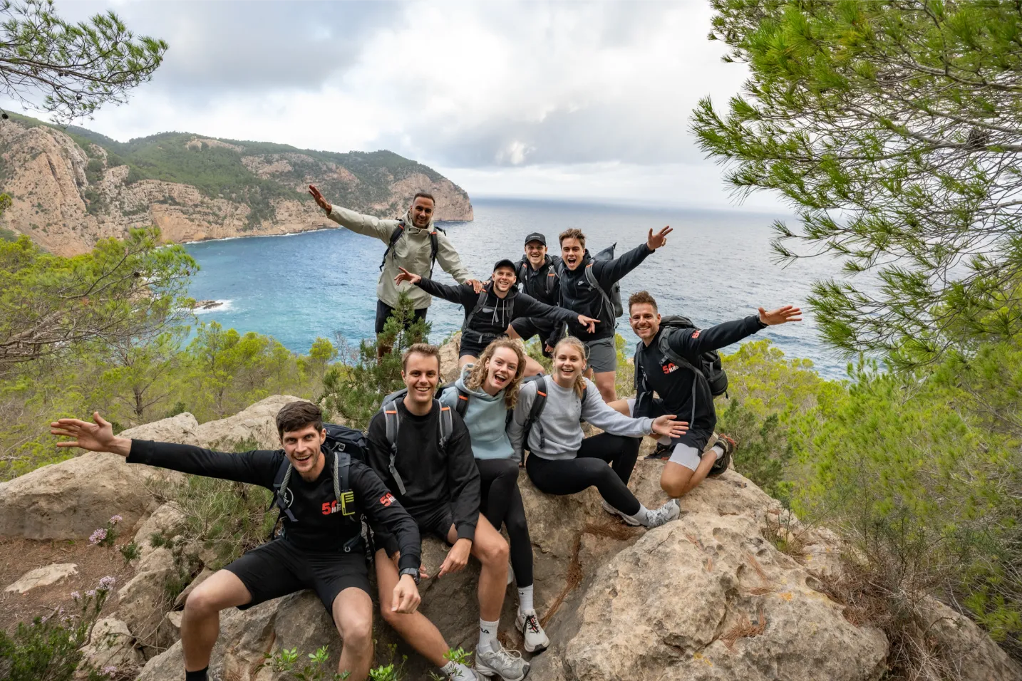 Negen jonge mensen zittend op een rots met rugzakken, sportieve kleding en armen omhoog langs kustlijn.