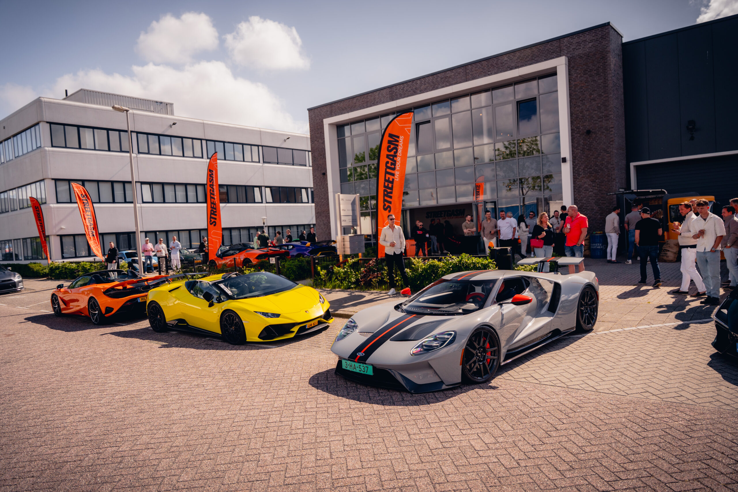 Luxe sportauto's in geel, oranje en zilvergrijs geparkeerd voor modern gebouw met oranje STREETGASM vlaggen