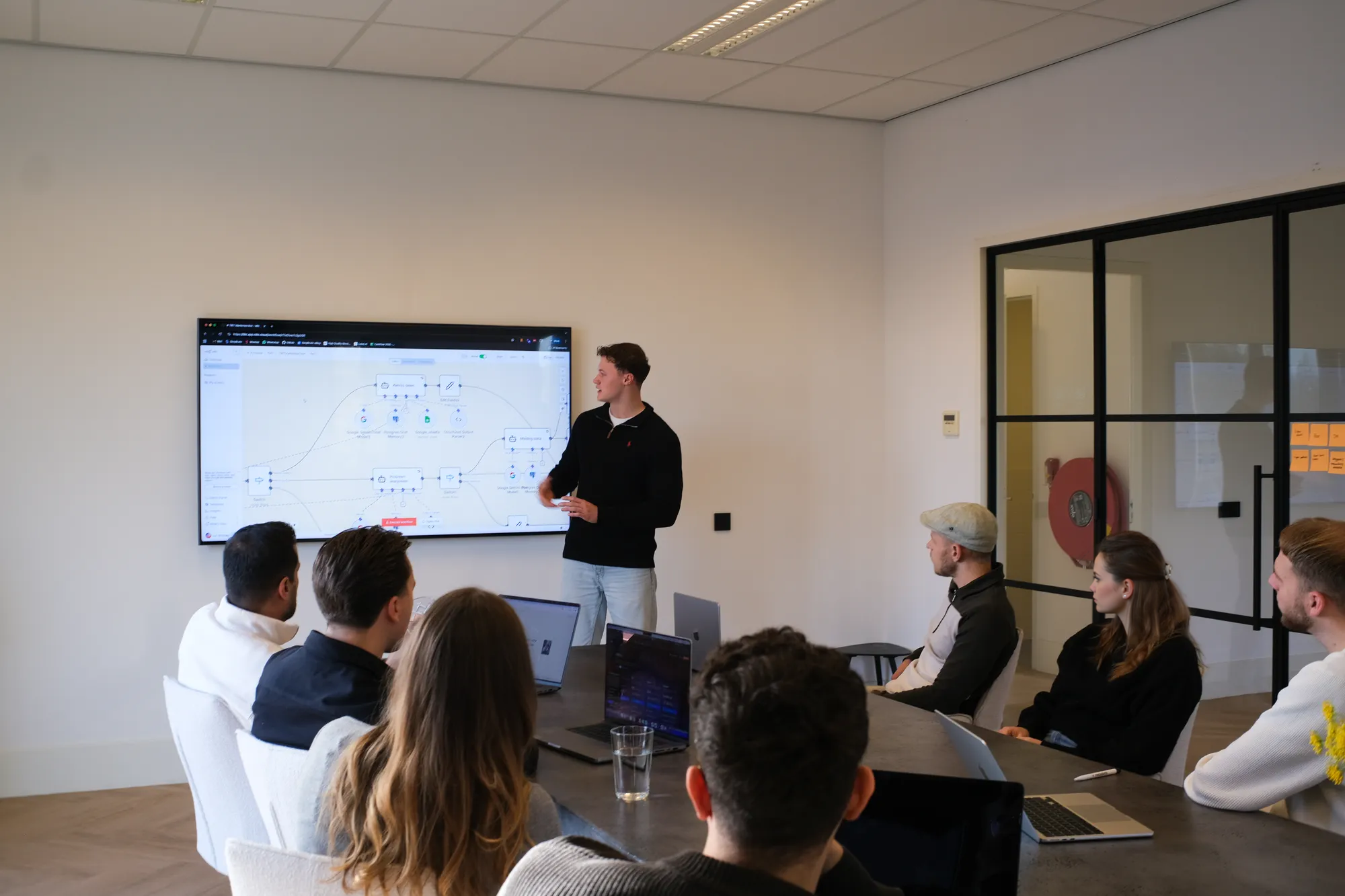 Mannen en vrouwen in vergaderruimte met laptops luisteren naar presentatie met diagrammen op groot scherm.